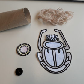 Ein aus Papier ausgeschnittener Skarabäus liegt auf einem Tisch neben einer Klorolle und einem Fadenknäuel