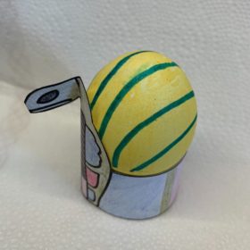 Auf einem weißen Hintergrund ein gelb-bkau gestreift bemaltes Osterei, das in einer gebastelten Krone sitzt.