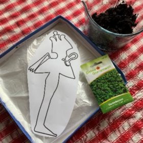 Auf einer rotkarierten Tischedecke liegt ein Deckel, in dem sich ein Umriss des Osiris befindet, Daneben steht ein Glasgefäß mit Erde. Auf dem Deckel liegt eine Packung Kresse.
