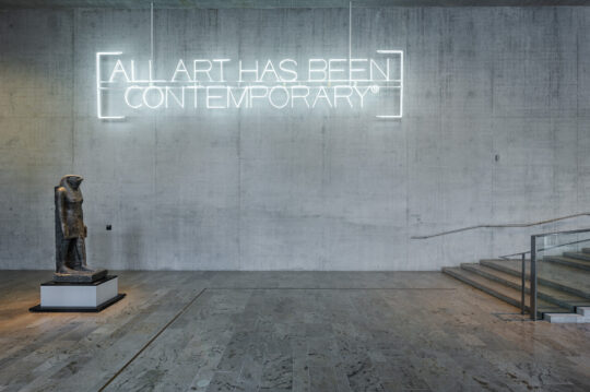 Auf einer hohen Betonwand hängt im oberen Bereich eine weiße Neoninstallation aus den Worten 