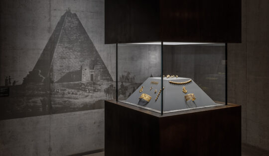 In einer viereckigen Vitrine ist Goldschmuck auf einem Pyramidenstumpf arrangiert. Dahinter eine Betonwand mit einem Bild der Pyramide der Amanishakheto nach einem Bronzestich