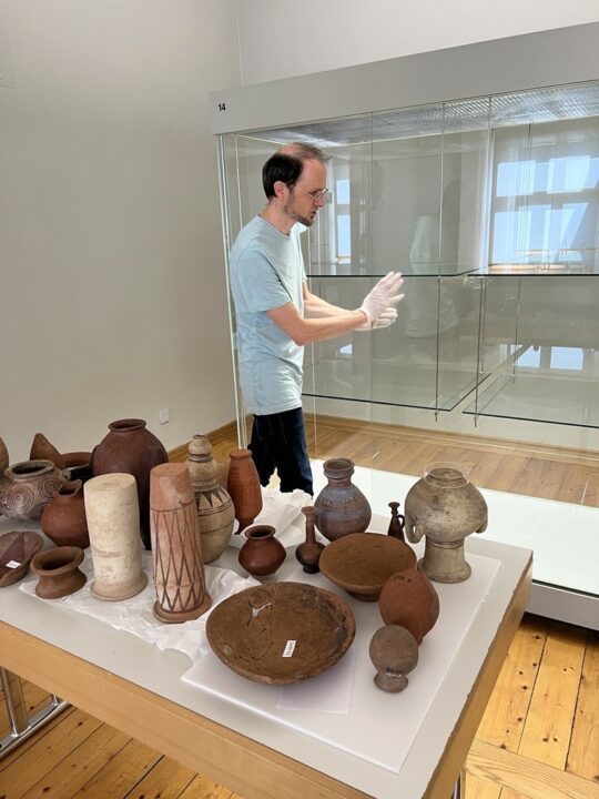 Kurator Jan Dahms steht hinter einem Tisch mit Keramikgefäßen. Hinter ihm eine leere Glasvitrine.