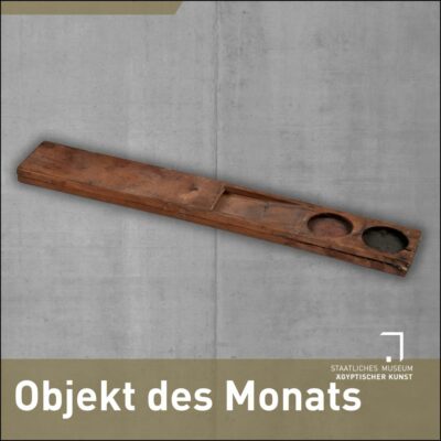 Eine Schreiberpalette aus Holz, längs-rechteckig, mit einer Vertiefung in der Mitte, um Binsen hineinzustecken und zwei runden Farbnäpfchen auf der rechten Seite, in denen noch Spuren von roter und schwarzer Farbe zu erkennen sind.