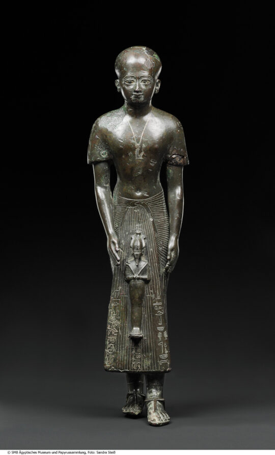 Bronzestatue eines stehenden, kahlköpfigen Mannes, der eine Statue des Osiris vor sich hält