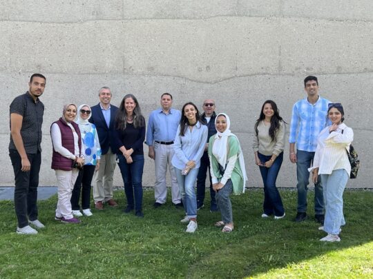 Eine Gruppe Menschen steht auf einer grünen Wiese vor einer Betonwand und lächelt freundlich in die Kamera