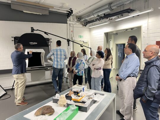 Einige Gruppe junge Menschen stehen in einem Fotoatelier. Im Vordergrund ein Tisch mit altägyptischen Objekten.