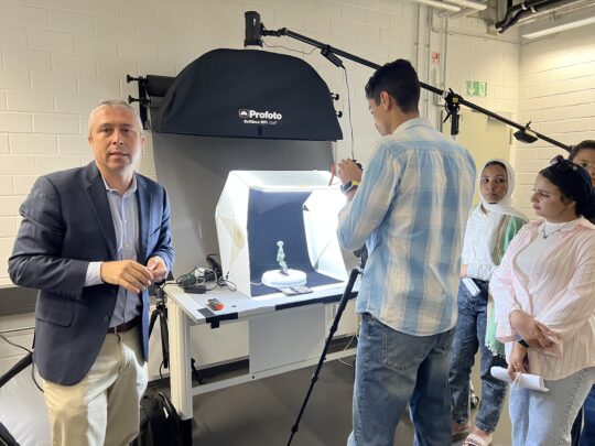 Einige Gruppe junge Menschen stehen in einem Fotoatelier. Auf einem Tisch steht hell erleuchtet eine altägyptische Statuette.