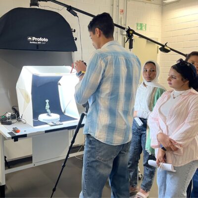 Einige Gruppe junge Menschen stehen in einem Fotoatelier. Auf einem Tisch steht hell erleuchtet eine altägyptische Statuette.