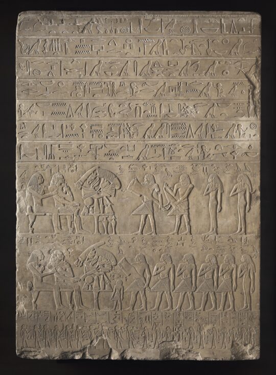 Eine altägyptische, hochrechteckige Stele aus hellem Stein. Die obere Hälfte ist on einer Hieroglypheninschrift bedeckt, in der unteren Hälfte finden sich zwei übereinanderstehende Bildfelder mit zahlreichen Personen, am unteren Ende der Stele eine Reihe mit sehr vielen klein dargestellten Personen.