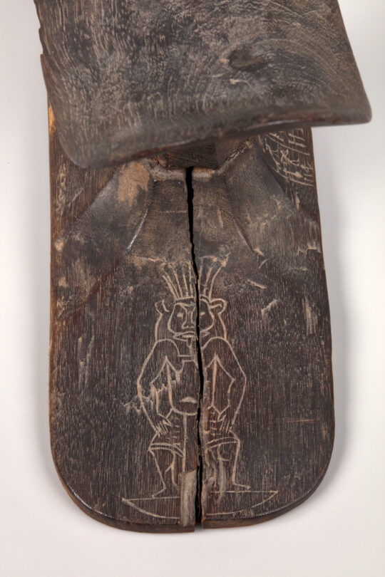Detailansicht des Fußes einer Kopfstütze aus dunklem Holz. In den Fuß ist eine Figur des Gottes Bes eingeritzt.